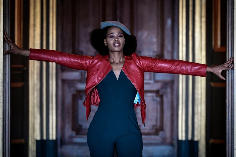 A woman with an Afro coming out from beneath a beret poses with her arms out in a gilded doorway, wearing a red leather jacket and a no-nonsense expression.
