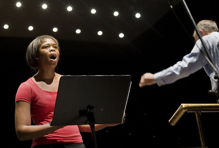 A woman in a red dress and short hair sings in fromt of a stand with music on it, an orchestra conductor visible to her side.