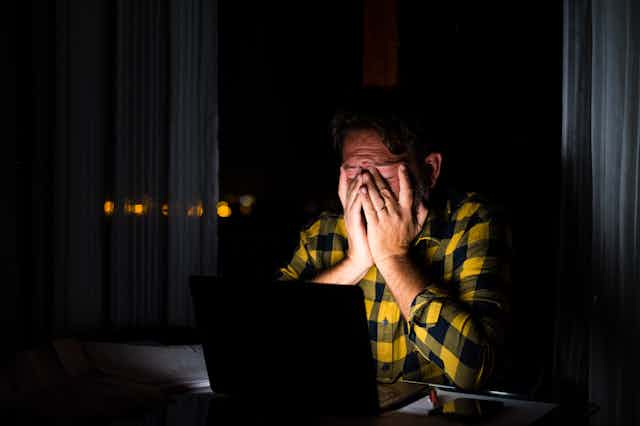 un hombre con camisa a cuadros negros y amarillos en una habitación a oscuras y con un ordenador que ilumina su cara, tapada parcialmente con las manos como gesto de cansancio.