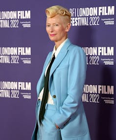 Tilda Swinton has her blonde hair in a tight curl and wears a baby blue suit.