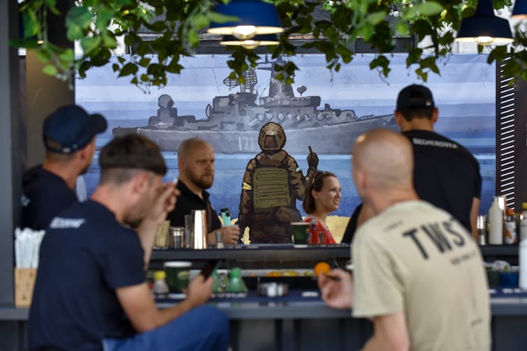 People in a Kyiv bar looking at a poster showing a defiant Ukrainian defender giving a Russian the finger.