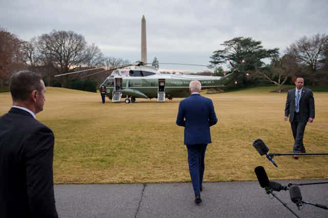 A man with white hair, in a blue suit, walks to a helicopter.