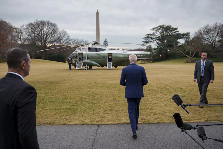 A man with white hair, in a blue suit, walks to a helicopter.