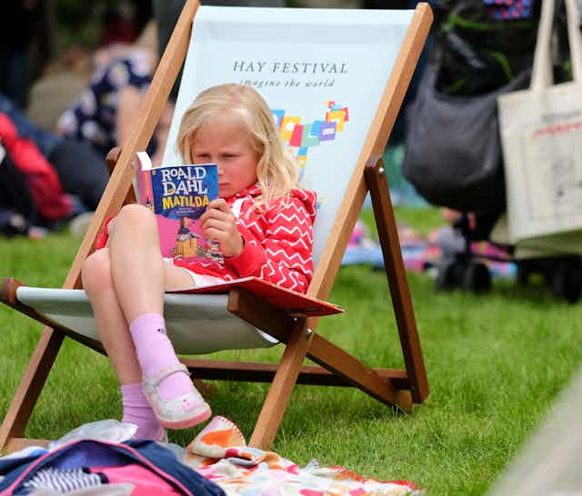 A young girl reads Matilda by Roald Dahl on a deck chair.