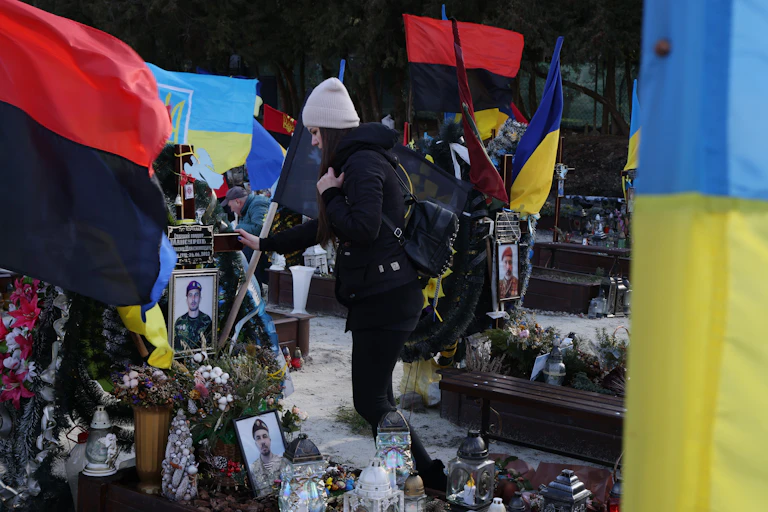 A woman wearing dark clothing and a light hat puts her hand on a grave that is covered in photos of a young man. Red and black flags and a blue and yellow flag surround her.