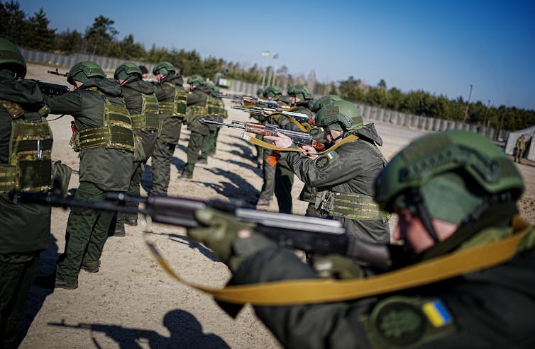 Two rows of soldiers in green outfits and helmets hold rifles all pointed in the same direction.