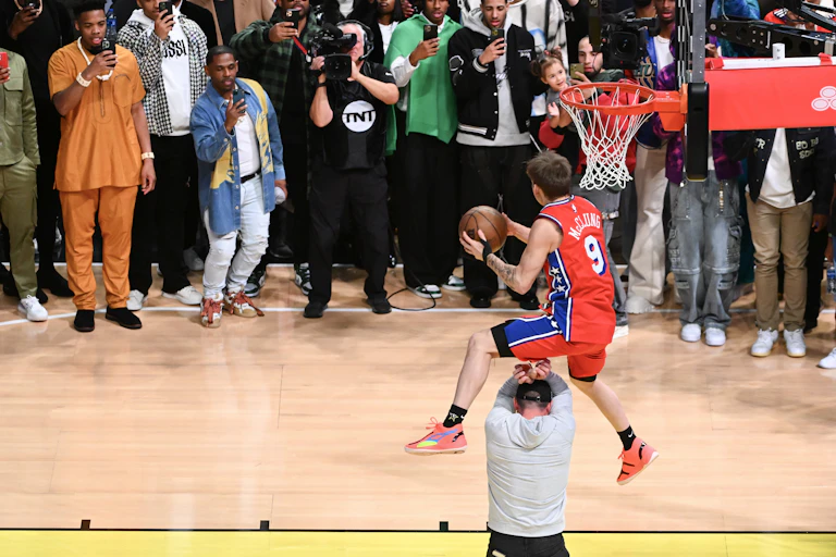 A man with a basketball jumps over another man to dunk the ball in a basketball hoop.
