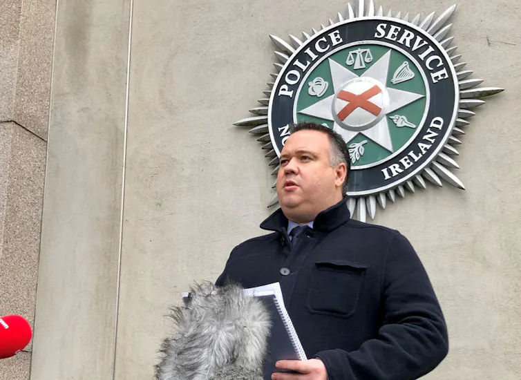 Man speaks into microphones below a large Police Service of Northern Ireland badge on the wall behind.