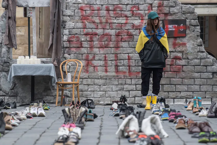 A woman wears a blue and yellow flag draped over her shoulders ad stands in front of rows of pairs of shoes.