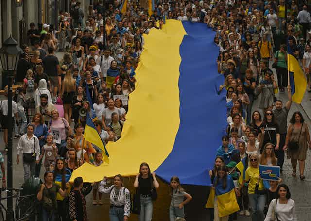 Hundreds of people carry a gigantic yellow and blue Ukrainian flag down a street.