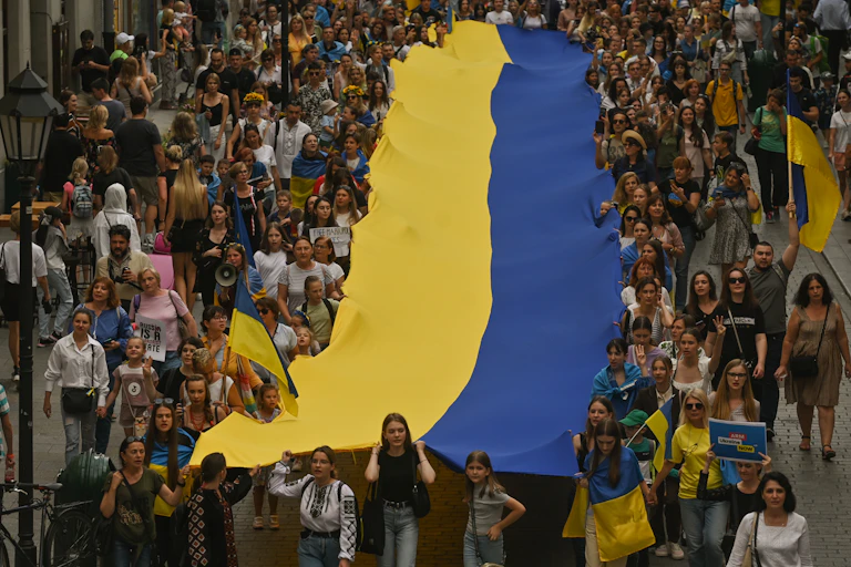 Hundreds of people carry a gigantic yellow and blue Ukrainian flag down a street.