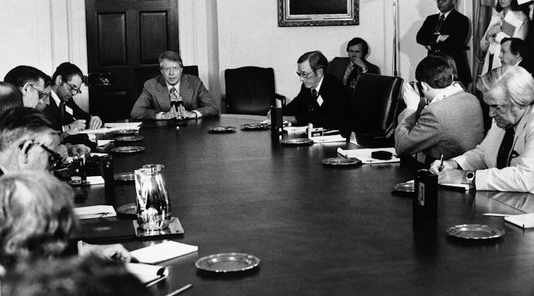 A number of people gather around a table, taking notes, as the person at the head of the table speaks.