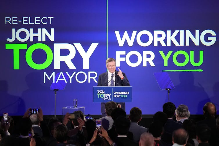 A man in a suit speaks from behind a podium. Behind him is a giant screen that says 'John Tory Mayor Working for You'