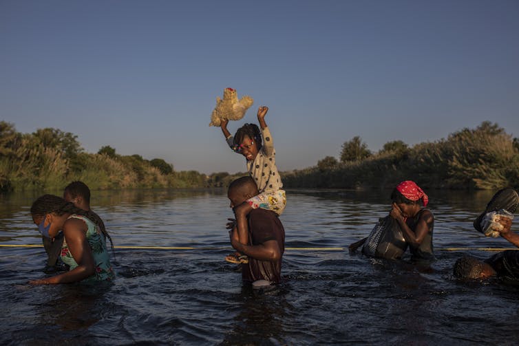 A girl holds her stuffed animal high above the water as migrants wade across a river.
