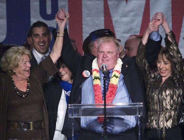 A man wearing a lei around his neck raises his arms in victory as he speaks from behind a podium. A small crowd of people stand behind him and two women on either side of him are also raising their arms