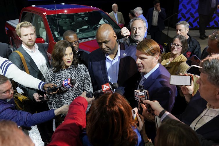 Three officials answer questions from reporters holding up microphones. A truck is in the background.