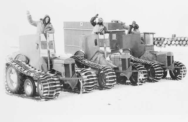 A black and white photo of 3 men waving from snow tractors.
