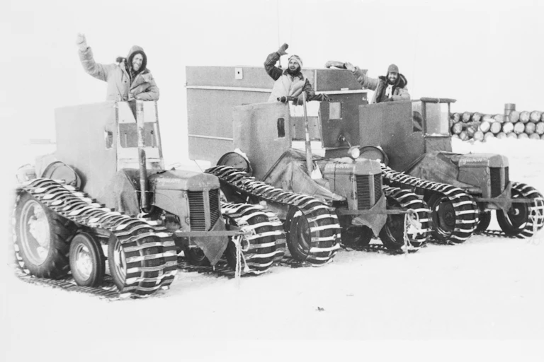 A black and white photo of 3 men waving from snow tractors.