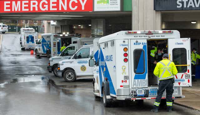 Paramedics at the open door of an ambulance in the foreground, with several other ambulances in the background under a red sign reading 'Emergency'