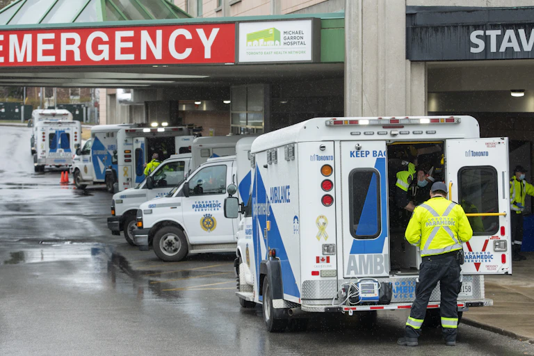 Paramedics at the open door of an ambulance in the foreground, with several other ambulances in the background under a red sign reading 'Emergency'