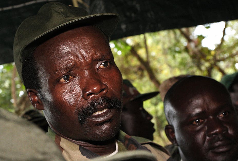A close-up shot of a man in a brown and green military uniform and green hat speaking. There are other men in the background dressed similarly.