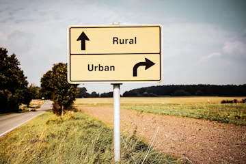 A road sign on the side of the road near a field. One arrow points towards "rural" and the other points towards "urban".