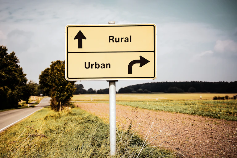 A road sign on the side of the road near a field. One arrow points towards "rural" and the other points towards "urban".