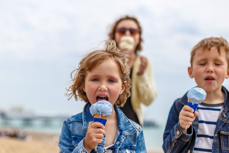Two children eating ice-creams with a woman in the background.