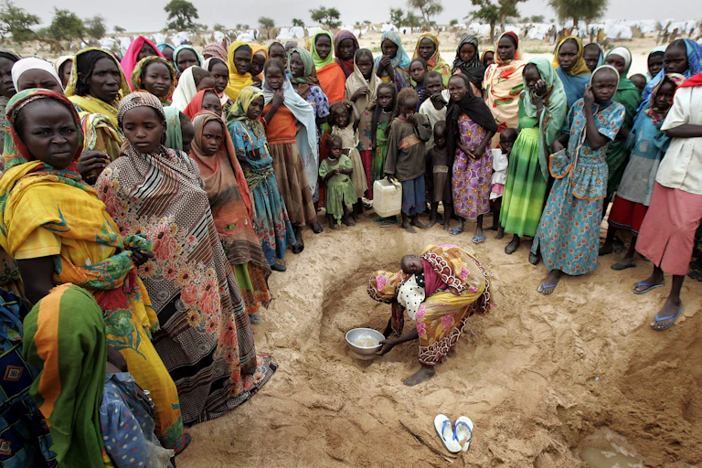 A group of people stand around a hole in the ground where one person squats with a bowl.