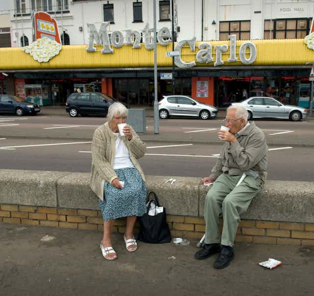 Two elderly people sit on a wall on a pavement drinking from white polystyrene cups.