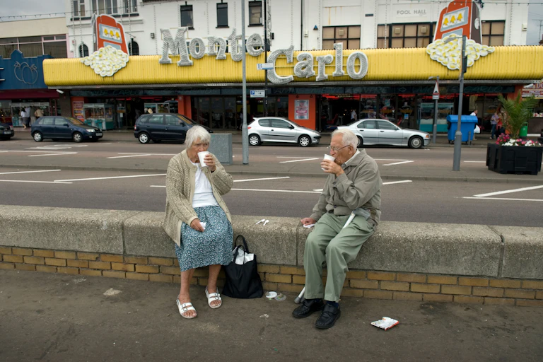 Two elderly people sit on a wall on a pavement drinking from white polystyrene cups.