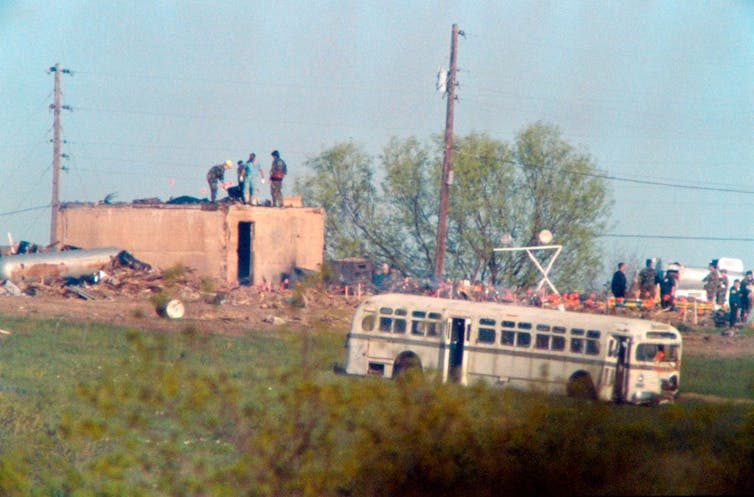 People search amid the remains of a burned building, with a school bus parked nearby.