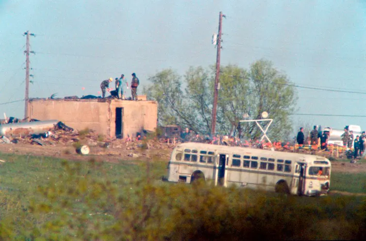 People search amid the remains of a burned building, with a school bus parked nearby.