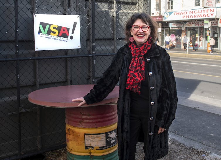 A woman smiling in front of a busy main road. She wears glasses, a red scarf and a black jacket.