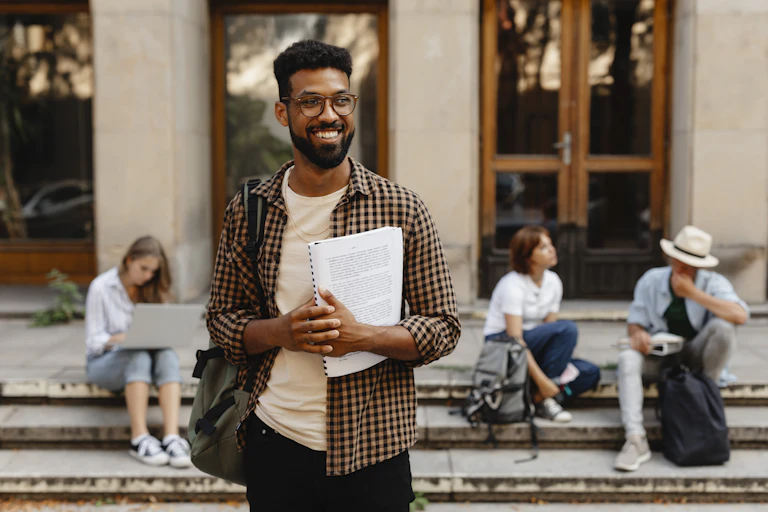 A smiling Black student holds a notebook and backpack in front of white students seated on the steps of a college.