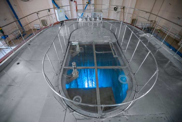 A concrete room with a circular hole in the middle surrounded with railings, with blue glowing water inside