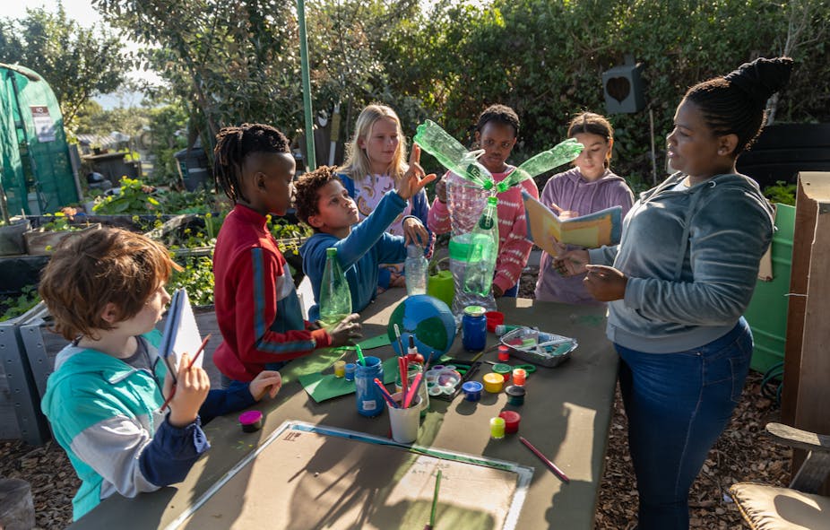 Six young children in casual clothes, some holding books and others paintbrushes, gather around a model of the earth and some plastic bottles fitted together while an adult, their teacher, talks to them