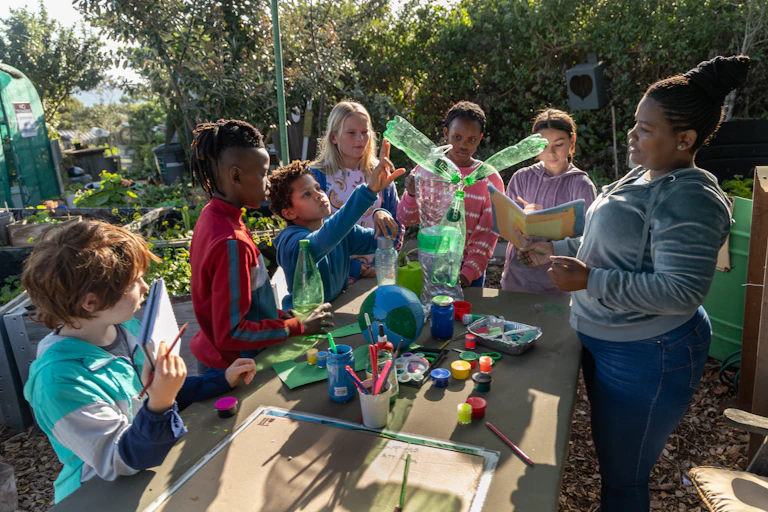 Six young children in casual clothes, some holding books and others paintbrushes, gather around a model of the earth and some plastic bottles fitted together while an adult, their teacher, talks to them