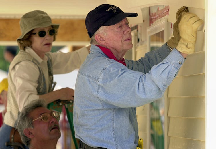 An elderly man and a woman attach siding to the front of a Habitat for Humanity home.