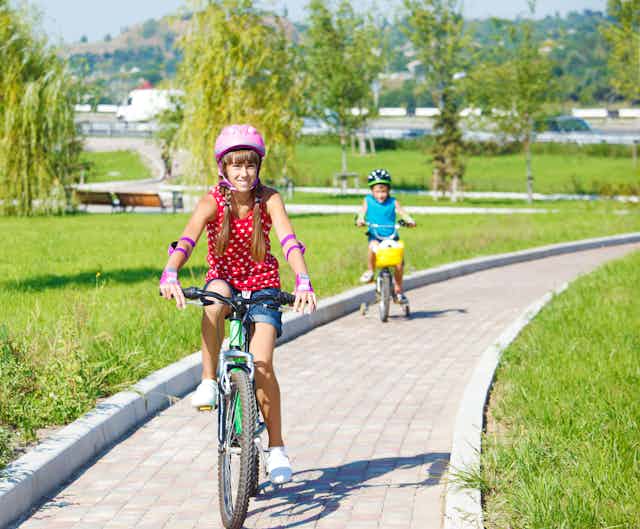 Una niña y un niño con casco y bicicleta atraviesan un parque. 
