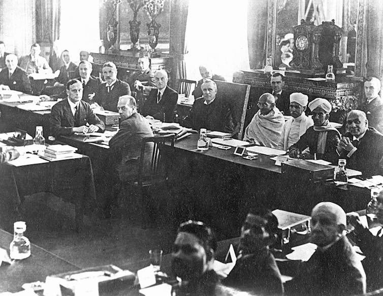 Black and white photo of formally dressed British and Indian men and Gandhi in his dhoti seated at long tables.