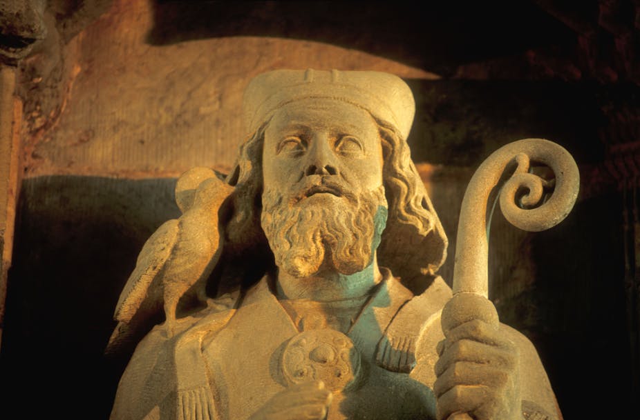 A stone statue of a bearded man wearing a hat. He has a dove on his right shoulder and is carrying a crozier in his left hand. 