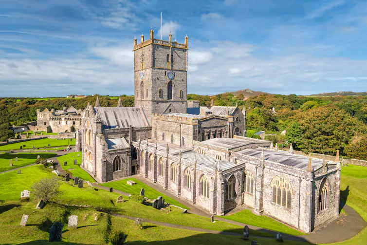 A very large medieval church sits within a wide expanse of green fields. The sky is blue.
