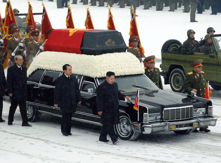 Men in suits and military uniforms walk alongside a hearse and army vehicles through the snow.