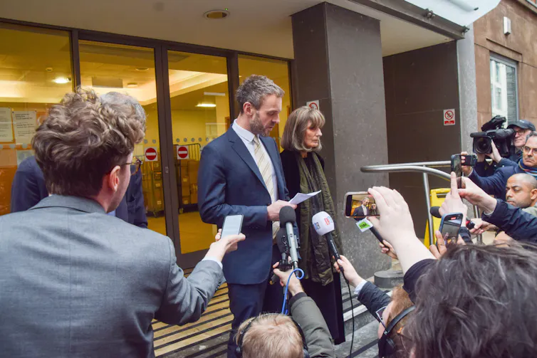 a man and woman read a statement while surrounded by members of the press extending microphones and recording devices