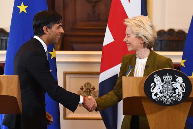 Rishi Sunak and Ursula von der Leyen smile and shake hands, both standing at podiums in front of an EU and UK flag