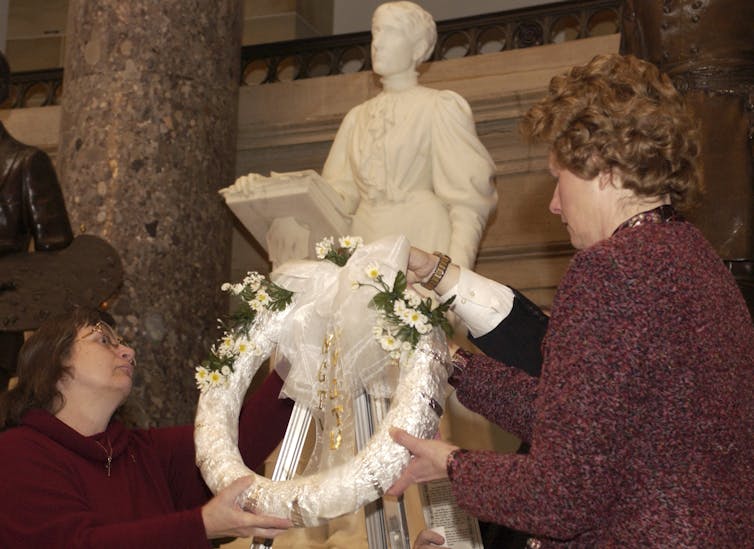 Two women lay a wreath at the statue of a woman holding a book in one hand.