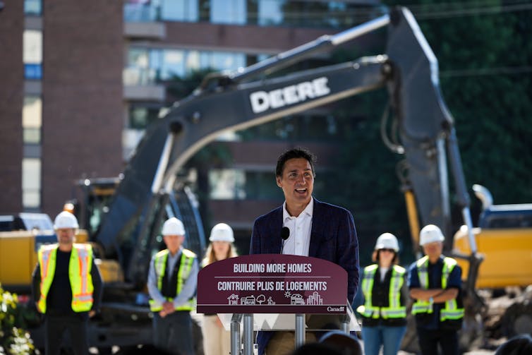 A man in a suit speaks from behind a podium that says 'Building More Homes' on the front of it. In the background a group of people wearing fluorescent vests and hard hats stand in front of an excavator.