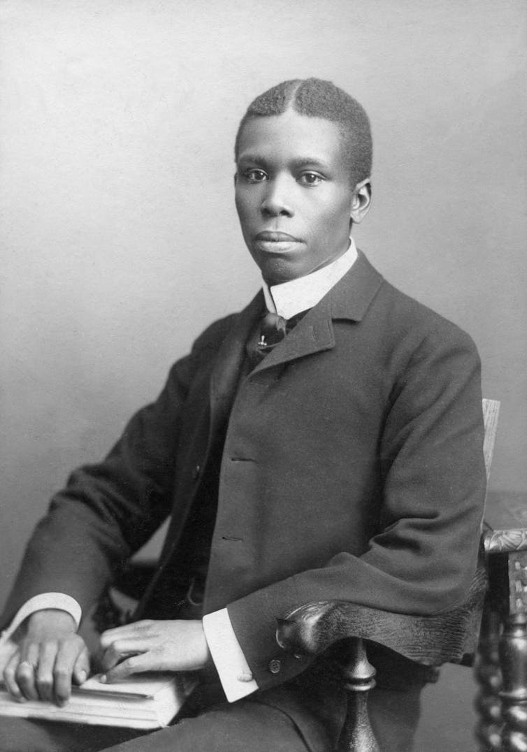 A young black man dressed a dark suit sits in a chair with a book and pen resting in his lap.