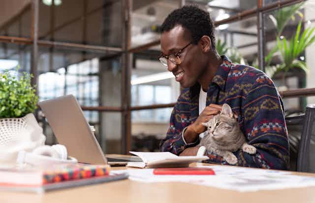 A man wearing glasses pets a cat while smiling into an open laptop.